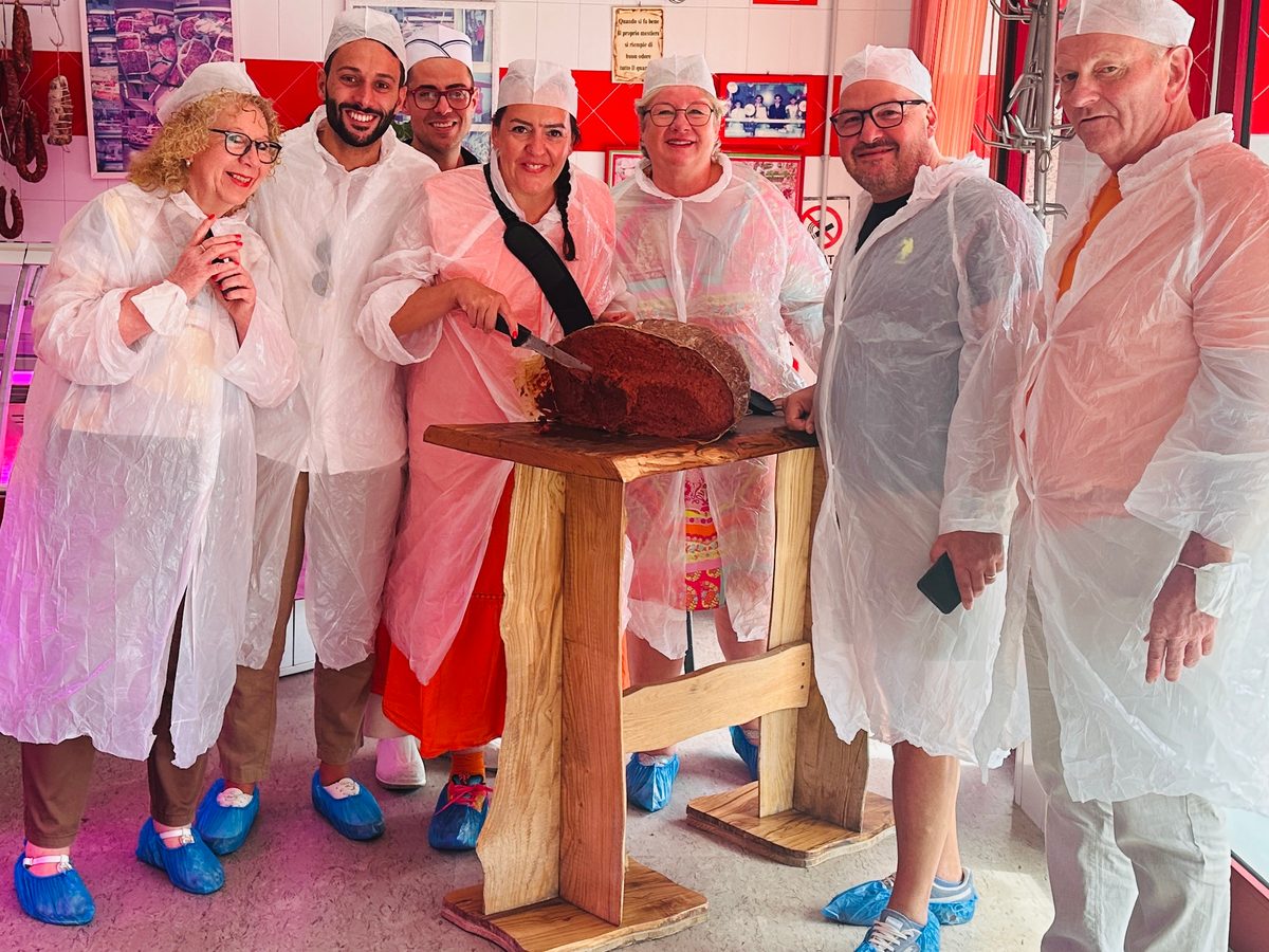 Unsere Gruppe in weißen Kitteln mit einer großen ’Nduja auf einem Holzbock im Verkaufsraum